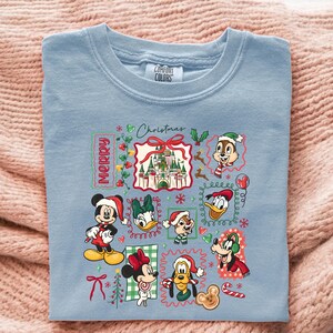 May include: Light blue Comfort Colors t-shirt featuring a Christmas-themed graphic. The design includes the word "Merry" and various Disney characters like Mickey Mouse, Minnie Mouse, Donald Duck, and Chip and Dale, all in festive attire.