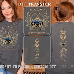 May include: Dark gray t-shirt featuring a graphic design with the text "VELARIS CITY OF STARLIGHT" and a mountain scene. The design includes gold and blue accents, with additional text below. The back of the shirt displays a moon phase design. The words "READY TO PRESS CUSTOM DTF" are also visible.