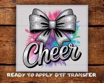 May include: A square design featuring a silver glitter bow with the word "Cheer" in a silver, cursive font. The background has a colorful watercolor splash of pink, purple, and teal. The text "READY TO APPLY DTF TRANSFER" is at the bottom.