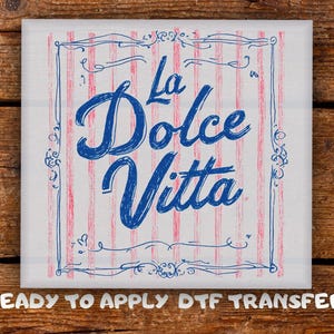 May include: Square white transfer with the words "La Dolce Vita" in blue script, framed by a blue decorative border. The background has vertical red and white stripes. The text "READY TO APPLY DTF TRANSFER" is at the bottom.
