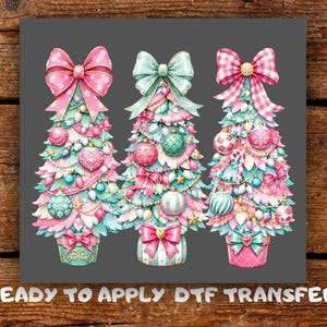 May include: A DTF transfer design featuring three stylized Christmas trees in shades of pink, mint green, and white. Each tree is decorated with ornaments, bows, and lights, topped with large bows. The text READY TO APPLY DTF TRANSFER is below.