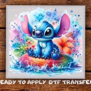 May include: A watercolor-style image of a blue cartoon character with large ears, sitting on a colorful seashell. The character is surrounded by splashes of blue, pink, and purple watercolor, with tropical flowers. Text at the bottom reads "READY TO APPLY DTF TRANSFER."