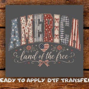 May include: A grey graphic with the word "AMERICA" in a patriotic design. The letters are filled with patterns like stars, floral prints, and stripes. Below, the phrase "land of the free" is written in cursive, with a heart-shaped flag.