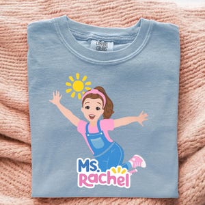May include: Light blue Comfort Colors crew neck t-shirt featuring a cartoon illustration of a girl with a sun graphic. The girl is wearing pink and blue clothing and the text "MS. Rachel" is below the illustration.