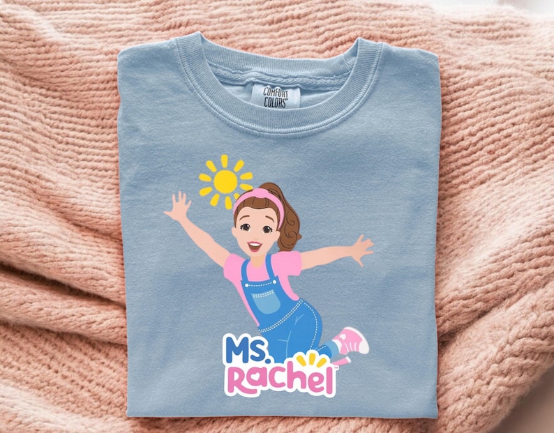 May include: Light blue Comfort Colors crew neck t-shirt featuring a cartoon illustration of a girl with a sun graphic. The girl is wearing pink and blue clothing and the text "MS. Rachel" is below the illustration.