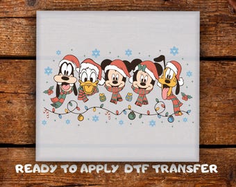May include: A light gray DTF transfer featuring cartoon characters Goofy, Donald Duck, Mickey Mouse, Minnie Mouse, and Pluto, all wearing Santa hats and scarves. They are surrounded by Christmas lights, snowflakes, and gifts. The text "READY TO APPLY DTF TRANSFER" is at the bottom.