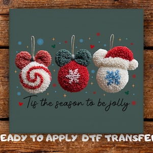 May include: Three Christmas ornaments with Mickey Mouse ears. The ornaments are red, white, and green with snowflake and swirl designs. The text "Tis the season to be jolly" is below the ornaments.