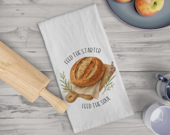 Op de afbeelding: Witte keukendoek met een aquarel illustratie van een brood op een houten snijplank, omringd door groen. De tekst "FEED THE STARTER FEED THE SOUL" is op de doek gedrukt.