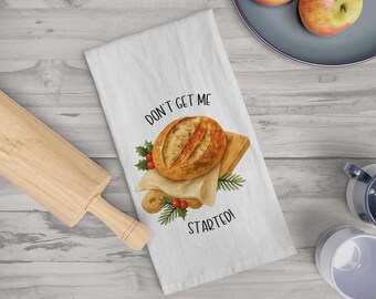 Op de afbeelding: Witte keukendoek met een illustratie van brood op een snijplank, met de tekst "DON'T GET ME STARTED!". Het ontwerp bevat hulst en rode bessen. Een houten deegroller en appels zijn ook zichtbaar.