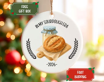 Puede incluir: Un adorno redondo de cerámica blanca con un cordón dorado para colgar. El diseño muestra una hogaza de pan de masa madre sobre una tabla de madera junto a un frasco con tapa de cuadros azules. El texto dice IN MY SOURDOUGH ERA y el año 2026.