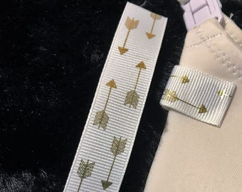 May include: A beige nursing bra strap featuring a white ribbon accent with a gold metallic arrow pattern. A matching white ribbon strip with the same gold arrow design is positioned next to the bra strap, showcasing the repeating arrow motif.