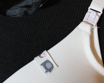 May include: A white nursing bra featuring a plastic clip on the shoulder strap and a small, light-colored fabric tag attached to the neckline. The tag displays a graphic of a gray elephant with a small pink accent. The bra is set against a black fabric.