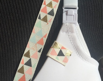 May include: A white nursing bra featuring a plastic clip on the strap and a decorative fabric strip with a geometric pattern of triangles in shades of coral, mint green, black, and metallic gold, matching a small fabric loop on the cup.