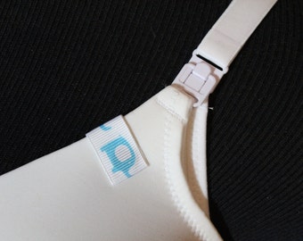 May include: A white nursing bra strap featuring a plastic adjustment clip and a small white fabric tag with a light blue elephant graphic attached to the upper edge of the cup, providing a clear view of the functional nursing clasp mechanism.
