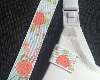 May include: A white nursing bra featuring a decorative ribbon strap with a floral pattern in coral, mint green, and gold metallic accents. The strap includes a white plastic nursing clip and a matching ribbon tab attached to the bra cup.