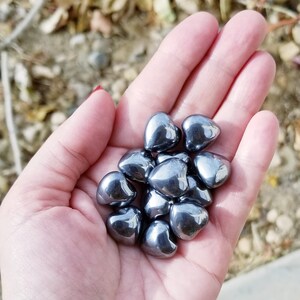 May include: A hand holding a collection of small, heart-shaped, dark gray hematite stones.