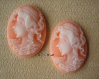 May include: Two oval-shaped cameo pendants with a white cameo image of a woman's face with detailed hair and a peach-colored background.
