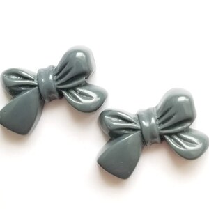 May include: Two gray plastic bow-shaped buttons. The bows are slightly raised and have a glossy finish.