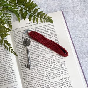 May include: A vintage key with a silver key ring and a burgundy crocheted key fob, lying on an open book. The book has text on the pages.