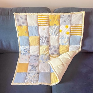 May include: A patchwork quilt in yellow, blue, and white. The quilt is made of squares with floral, striped, and solid patterns. Two yellow sun-shaped buttons are sewn onto one of the squares. The quilt is approximately 100 cm by 127 cm.