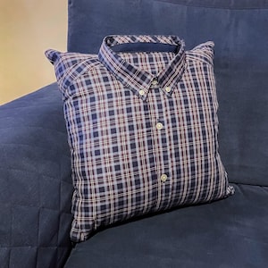 May include: A cushion designed to resemble a button-down shirt. It has a blue, white, and burgundy plaid pattern. The collar and button details are visible. The cushion is placed on a blue sofa.