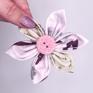 May include: A pink fabric flower with a pink button in the centre. The flower has a white background with a floral pattern and a grey and purple pattern on the back.