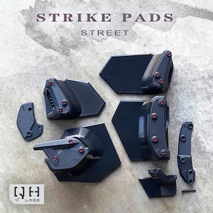 May include: An assortment of black strike pads with red accents and hardware, arranged on a gray surface. The text "STRIKE PADS STREET" is displayed at the top. The image includes various shapes and sizes of pads, suggesting a protective gear set.