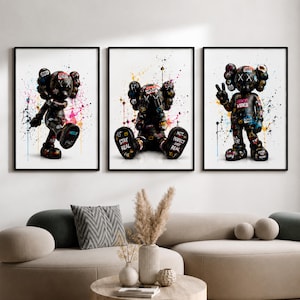 Peut inclure: Trois impressions d'art mural encadrées représentant des personnages noirs de style dessin animé avec des yeux en forme de X, couverts d'éclaboussures de peinture colorée et de textes de style graffiti comme "STAY REAL" sur fond blanc.