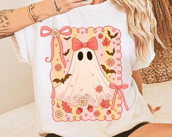 May include: White t-shirt featuring a cute ghost design with a pink bow, bats, and floral accents. The ghost is draped in a white sheet with eye holes. The design is framed with a pink border and ribbons, perfect for Halloween.