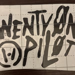 May include: Black vinyl sticker with the words "twenty one pilots" in a stylized font. The band's logo, a circle with two vertical lines, is also included. The sticker is on a white grid background.