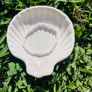 Pode incluir: Um prato branco iridescente em forma de concha com uma pulseira branca com contas no interior. O prato tem um brilho perolado e está colocado sobre um fundo de relva verde, criando uma exibição natural e elegante.