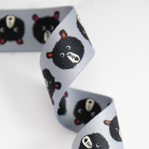 May include: A light gray ribbon with a repeating pattern of black bear faces. The bears have white muzzles, small red ears, and black eyes. The ribbon is curled and appears to be made of a smooth, satin-like material.