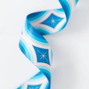 May include: A close-up of a blue and white ribbon with a repeating geometric pattern. The ribbon features a series of blue and light pink curved stripes, with diamond shapes containing a star design. The ribbon is curled and twisted.
