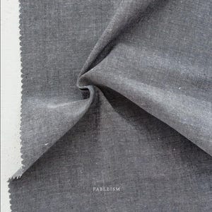 May include: Close-up of a gray fabric sample with a textured weave. The fabric is folded, revealing its depth and texture. The word "FABLEISM" is printed in white at the bottom right corner. The fabric's edge has a zigzag pattern.