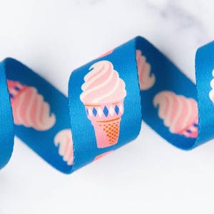 May include: A blue ribbon with a repeating pattern of pink ice cream cones. The ice cream cones have a pink swirl and a pink and orange cone with blue diamond accents. The ribbon is curled and has a white background.