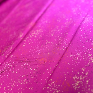 May include: A close-up of a vibrant fuchsia fabric with a speckled pattern. The fabric has a soft sheen and is folded, creating diagonal lines across the image. The speckles are in shades of white, orange, and purple.