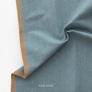 May include: A piece of blue fabric featuring a repeating pattern of small white crosses. The fabric has a solid tan border along one edge and the word FABLEISM is printed in white near the bottom center of the textile material.