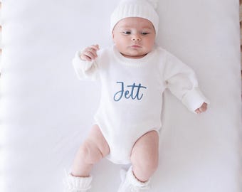 May include: A white long-sleeve knit bodysuit featuring the name Jett embroidered in blue script across the chest. The outfit includes a matching white knit beanie with a pom-pom on top and a pair of white knit booties with ribbon ties.