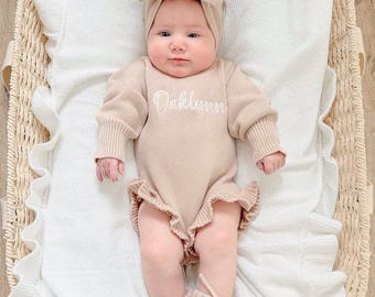 May include: A beige long-sleeved knit romper featuring a ruffled hem and the name Oaklynn embroidered in white script across the chest. The outfit includes a matching beige knit headband with a large bow and a pair of beige knit booties.