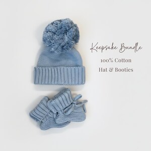 May include: A light blue knit hat with a large pom-pom and a pair of matching knit booties with tie closures. The text "Keepsake Bundle 100% Cotton Hat & Booties" is printed to the right of the items, which are made of 100% cotton material.