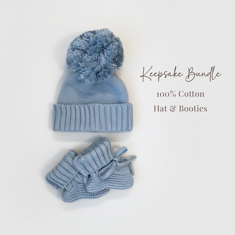 May include: A light blue knit hat with a large pom-pom and a pair of matching knit booties with tie closures. The text "Keepsake Bundle 100% Cotton Hat & Booties" is printed to the right of the items, which are made of 100% cotton material.