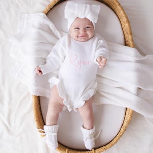 May include: A white knitted long-sleeve romper with ruffled edges and the name Ryver embroidered in pink script on the chest. The outfit includes a matching white knitted headband with a large bow and white knitted booties with tie closures.