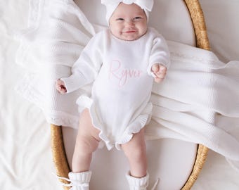 May include: A white knitted long-sleeve romper with ruffled edges and the name Ryver embroidered in pink script on the chest. The outfit includes a matching white knitted headband with a large bow and white knitted booties with tie closures.