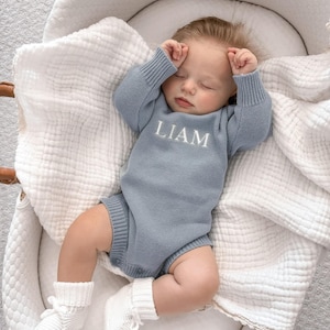 May include: A dusty blue long-sleeved knitted baby romper featuring the name LIAM embroidered in white thread across the chest. The garment has ribbed cuffs at the wrists and legs, and is worn by a sleeping infant resting on a white muslin blanket.