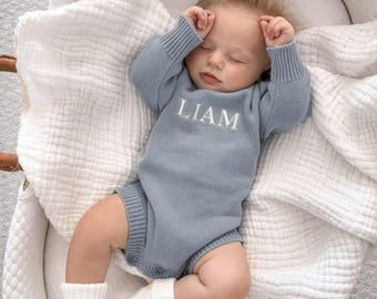 May include: A dusty blue long-sleeved knitted baby romper featuring the name LIAM embroidered in white thread across the chest. The garment has ribbed cuffs at the wrists and legs, and is worn by a sleeping infant resting on a white muslin blanket.