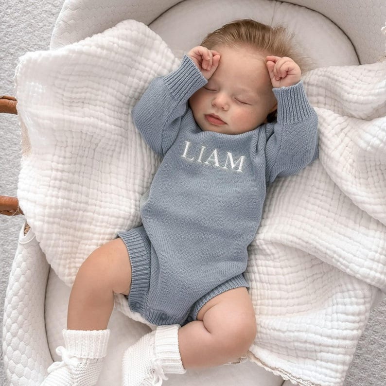 May include: A dusty blue long-sleeved knitted baby romper featuring the name LIAM embroidered in white thread across the chest. The garment has ribbed cuffs at the wrists and legs, and is worn by a sleeping infant resting on a white muslin blanket.