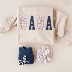 May include: A beige sweatshirt with the word MAMA embroidered in alternating solid navy blue and floral patterned block letters. The sleeve features embroidered names and a small blue flower. Below are a navy blue wrap shirt and a floral shirt.