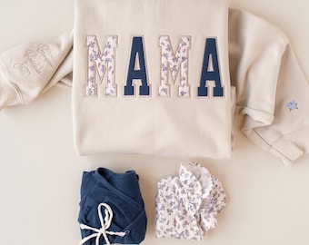 Può includere: Una felpa beige con la parola MAMA ricamata in lettere maiuscole alternate tra blu navy tinta unita e un motivo floreale. La manica presenta nomi ricamati e un piccolo fiore blu. Sotto ci sono una maglia a portafoglio blu navy e una maglia a fiori.
