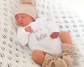 Può includere: Un body bianco a maniche lunghe con la scritta he's here ricamata in marrone chiaro, abbinato a un berretto in maglia marrone chiaro coordinato con un grande pon pon e un paio di scarpine in maglia marrone chiaro con lacci per neonato.