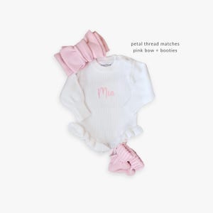Pu&ograve; includere: Un body in maglia bianco a maniche lunghe con orlo a volant e il nome Mia ricamato in filo rosa sul petto. Il set include una fascia con fiocco a coste rosa e un paio di scarpine in maglia rosa. Testo: petal thread matches pink bow + booties.